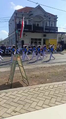 A marching band in uniform walks in formation down a street. They are wearing blue jackets with gold trim and white pants. Some members carry batons. They are walking past a building with a red tile roof and balconies. There are people on the sidewalk watching the parade. An A-frame sign is in the foreground.