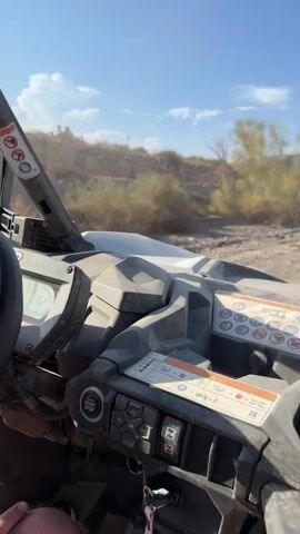 This is a first-person perspective video from a UTV (Utility Task Vehicle) driving on a dusty dirt road. The scene is outdoors in a bright, sunny environment, suggesting it's daytime with clear skies and warm weather. The setting appears to be a desert or arid region, with mountains visible in the background and sparse vegetation along the roadside. The UTV is moving forward, kicking up dust from the road.

In the foreground, the dashboard of the UTV is visible, cluttered with various controls and warning labels. One prominent label features several circular icons with red borders and diagonal lines, indicating prohibitions or warnings, such as "no smoking" and symbols related to personal protective equipment. Another label shows a gear selection indicator and other operational symbols. A key is visible in an ignition slot.

The activity taking place is recreational driving in an off-road vehicle. The UTV in front, which the camera is following, is also kicking up a significant amount of dust, emphasizing the dry and dusty conditions. A side mirror on the UTV reflects an image of a person, likely the passenger, holding a smartphone and wearing headphones. This reflection offers a glimpse of the occupants and suggests they are enjoying an excursion.

The overall mood conveyed is one of adventure and outdoor exploration. The bright sunlight and open landscape contribute to a sense of freedom and excitement. The location cues, like the arid terrain and mountains, combined with the provided context of Pechina, Spain, situate the activity in a specific geographical area known for such landscapes.
