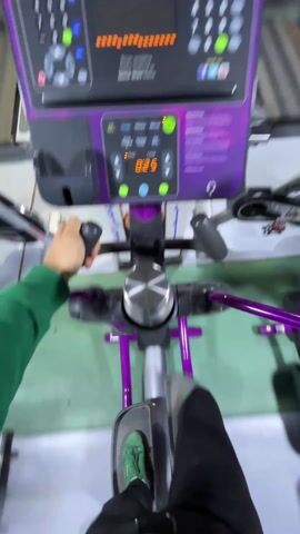 A first-person perspective video shows a person using an elliptical machine at a gym. The machine is purple and black, with a digital display showing the distance and level. The person is wearing a green sweatshirt and black pants with green sneakers. They grip the machine's handles and move their legs in a circular motion. In the background, other exercise equipment and people can be seen. A large advertisement for Planet Fitness is visible above the elliptical.