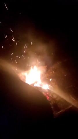 The video shows a small bonfire burning in the dark. The fire is contained in a square pit. Embers glow red and orange at the base of the fire, with brighter yellow and white flames rising above them. Smoke rises from the fire and drifts upward. A few sparks fly into the air. The edges of the frame are dark, suggesting the nighttime setting. A small portion of an arm or leg can be seen in the bottom left corner of the frame.