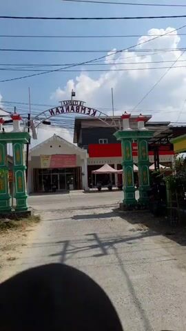 The video begins by showing an archway with text that appears to read "KECAMATAN KEMBANGAN". A street is visible beneath the archway, with several buildings on either side. One building has a red awning and the text "YU-WINFLY" on it. Another building has a white facade and a red sign with a white logo. Tents are set up in front of the Yu-Winfly building, with motorcycles parked underneath them.

As the video progresses, the camera pans to the right, revealing a cell tower in the distance. More buildings are visible along the street, including what appears to be a food stall. Two motorcycles are seen riding on the street, each with at least one person on it. The overall environment appears to be a street in a town or city, with a clear sky and some clouds. The video seems to be shot from a moving vehicle, likely a motorcycle, judging by the perspective.