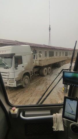 The video is shot from inside the cabin of a crane, looking out through the front windshield. The view is angled slightly downwards and to the left, showing an industrial or construction site.

Dominating the external view is a large, white semi-trailer truck with a long, flatbed trailer. The truck is parked on a muddy, dirt-covered surface. Its wheels are partially obscured by mud. The trailer is loaded with what appear to be several long, rectangular pre-fabricated sections or beams, stacked horizontally.

In the upper left portion of the external view, a crane hook with attached chains is visible, hanging in mid-air, positioned above the truck's trailer. This suggests the crane is either preparing to lift items from the truck or lower items onto it.

In the background, running parallel to the truck, is a long, low-rise building with multiple windows. The building appears to be made of light-colored, possibly concrete or pre-fabricated, panels.

Inside the crane cabin, several operational elements are visible. On the upper left, mounted above the windshield, is a display screen showing a detailed graphic or possibly a camera feed. To its right, another display screen is visible, showing a graphic representation of crane operations, likely a load chart or radius indicator, with green lines and data. A red emergency stop button is prominent on the right side of the cabin interior, below a black handle or lever. A crumpled white tissue or cloth is tucked into a crevice to the right of the rightmost display screen.

The weather appears overcast, and there are small water droplets on the windshield, indicating it might be raining or has recently rained. The ground is clearly muddy, consistent with a construction or heavy industrial environment. The overall scene depicts a cargo loading or unloading operation at a work site during the daytime.
