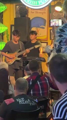 A band performs on a stage in an Irish pub. Two young men are playing guitars, and a young woman is singing. Several audience members are visible in the foreground, some recording the performance on their phones. The stage is decorated with green lanterns and a Guinness advertisement. The overall atmosphere is lively and casual.