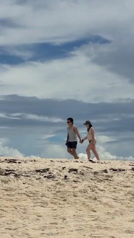A couple walks hand-in-hand across a sandy beach. The man is wearing a gray tank top, dark shorts, and sunglasses. The woman is wearing a light-colored top, a dark-colored baseball cap, and bikini bottoms. They are walking from left to right across the frame. The beach is covered in sand and some debris. The sky above them is blue with scattered clouds.
