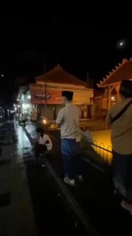 At night, a group of men are seen strolling in a brightly lit outdoor area featuring traditional architecture. The men are wearing traditional Muslim attire, including sarongs and peci caps. One man is wearing a light blue shirt and jeans, while another wears a brown shirt and a sarong. A mosque with a dome and minarets stands in the background, illuminated by lights.  The men are walking along a path bordered by a low fence, with a traditional tower-like structure visible nearby.  The scene evokes a sense of cultural exploration or religious observance. The video appears to have been taken handheld and possibly by one of the men who are walking and talking in the video.