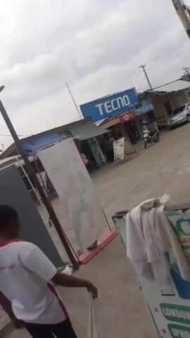 The video begins with a close-up shot of an "Ace Gadgets" sign, advertising "London Used Phones," "iPhones Available," and "Phone Swapping." The camera pans up to reveal a street scene with a "Tecno" sign in the background. A person wearing a white and red shirt, black pants, and sandals walks into the frame. They are seen holding a white cloth, possibly a towel. The camera focuses on the person's feet and the concrete ground, which appears damp with water stains.