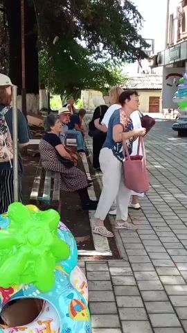 The video shows a group of people in an outdoor setting, possibly a park or a street. In the foreground, there are several inflatable objects, including a green one shaped like a sea creature and a ring with colorful donuts. The middle ground features a wooden bench with an elderly woman sitting on it, wearing a patterned dress and holding a checkered bag. Next to her, a man in a hat is seated. To the right, a woman in a blue and white patterned top and white pants is standing, holding a pink bag with a white star. She appears to be interacting with another woman in a white top and dark skirt. Further back, more people are visible, some walking and others sitting. The environment includes trees with visible trunks and leaves, a paved walkway, and a building in the background. The overall lighting suggests it is daytime.