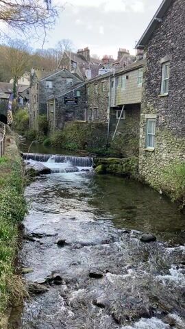 A short video shows a narrow, gently flowing river running through a quaint, old town. The buildings on either side are made of gray stone, with some featuring multiple stories and window shutters. A small waterfall is visible in the middle of the river.  The sky is overcast. There are no people visible in the video. The river flows over rocks and pebbles, and there is vegetation along the banks. The overall scene evokes a sense of peacefulness.