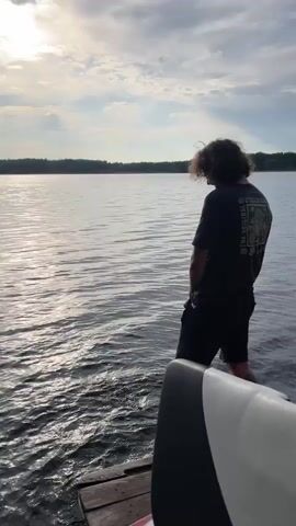 The video shows a man in a dark t-shirt and dark shorts standing at the edge of a lake. He appears to be urinating into the water. After finishing, he turns to face the camera and smiles. The sky is overcast, but there is bright sunlight breaking through. The lake is surrounded by trees in the distance. The camera appears to be on a boat docked at a small wooden pier.