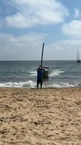 The video shows a person maneuvering a motorboat onto a sandy beach. The individual, wearing a blue shirt, guides the boat from the shoreline as it approaches the beach. The boat, which appears slightly weathered, has a canopy structure. The waves of the ocean create white foam as the boat gets closer to the shore. The beach itself is a light tan color, and the sky is blue with scattered clouds. In the distance, there's a sailboat visible on the water.
