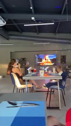 The media shows two people, a woman and a boy, sitting at a table in a dimly lit room with disco lights projected onto the ceiling. A large screen in front of them displays a karaoke video with Russian lyrics. The woman is holding a microphone and appears to be singing along. The boy is looking at the screen. There are drinks on the table, suggesting they are at a restaurant or bar. The scene appears to be set in the evening or indoors. The visible text on the screen is in Russian and appears to be song lyrics.