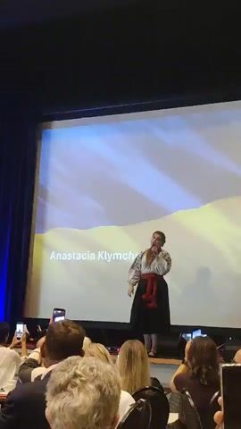 A woman named Anastacia Klymchuk stands on a stage, singing into a microphone. She is wearing a traditional Ukrainian outfit, consisting of an embroidered white shirt with red detailing, and a black skirt with a red sash. Behind her, a large screen displays the Ukrainian flag, with a yellow and blue design. The audience, composed of people of various ages, watches her performance, with many holding up their phones to record. The lighting on stage is dim, while the screen behind the performer is brightly illuminated. The overall environment appears to be an indoor venue, possibly a conference hall or auditorium, with dark curtains visible on the sides of the stage.