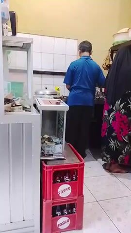 In the video, two individuals, an adult male and an adult female, are seen in what appears to be a kitchen or food preparation area. The male, wearing a blue shirt and dark pants, is standing and facing away from the camera, appearing to be engaged in cooking or food preparation. The female, wearing a black headscarf and a long dress with a floral pattern, is also standing and facing away from the camera, positioned to the right of the male, also seemingly involved in food preparation.

The environment shows tiled walls and a tiled floor. In the foreground, a red plastic crate is stacked with bottles, one of which has a "SOSRO" label visible. Adjacent to the crate, there is a display case or counter with items inside, including what appears to be money and other small objects. A metal structure with shelves is visible behind the male, and a dark wooden cabinet with shelves and various items is behind the female. The lighting suggests an indoor setting. The actions depicted are minimal, primarily standing and attending to tasks at a counter or stove.