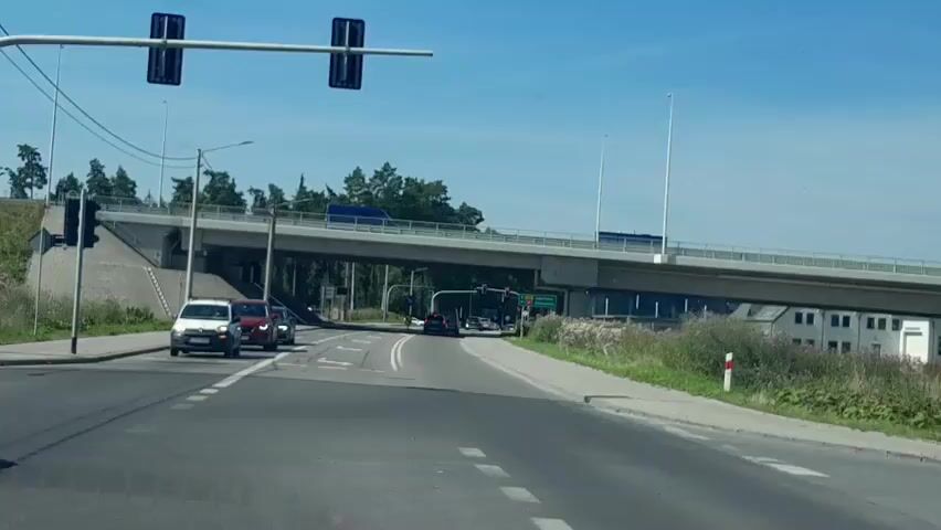 The video opens with a view of a street intersection with traffic lights. Several cars are stopped at the intersection, and a large overpass is visible in the background. The camera then pans to the right, showing a grassy embankment with trees and poles on top. Another car drives by on the overpass. The video continues to pan to the right, revealing more of the embankment and trees. The scene is bathed in sunlight, and the sky is clear and blue.