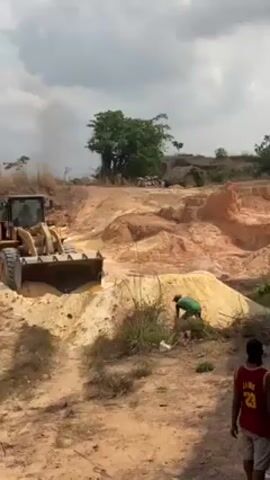 The scene unfolds in a dirt-filled environment, suggesting a construction or excavation site. A yellow, heavy-duty front-end loader dominates the view, positioned on a mound of sandy soil. A man in a green shirt stands beside the loader, shoveling sand, while another man wearing a red and black jersey is a short distance away, seemingly observing. The landscape is characterized by undulating sand hills and sparse vegetation, including trees and shrubs. In the background, a rudimentary structure with a thatched roof is visible, and smoke rises from an area near the horizon, creating a slightly hazy atmosphere.  The sky is overcast with heavy cloud cover.