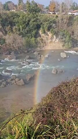The video shows a waterfall cascading into a rocky riverbed. At the beginning of the video, a rainbow is visible in the mist created by the waterfall. The water is clear and fast-flowing, rushing over the rocks. The surrounding environment includes lush vegetation and trees. The sky is clear with some scattered clouds. The camera pans up to reveal the full expanse of the waterfall. The scene is outdoors and appears to be a natural environment.