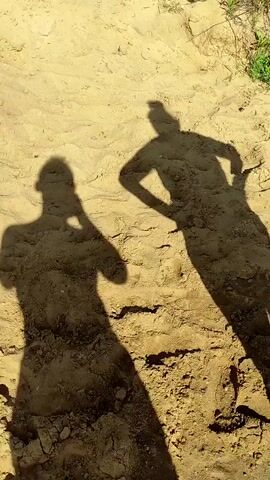 Two human shadows are cast on sandy ground. The shadow on the left appears to be holding a phone to its ear. The shadow on the right has its hands on its hips. Green foliage is visible in the upper right corner of the frame.