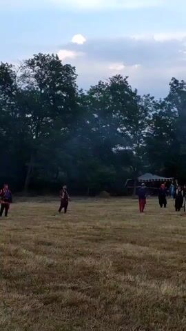 A wide shot of a historical reenactment shows a large group of people dressed in historical clothing engaged in a mock battle in an open field. Several individuals fire muskets, producing large puffs of white smoke. The ground is covered in dry, golden grass, and a line of dark green trees forms the background. The sky is partly cloudy with some visible blue sky.