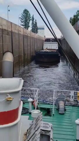 The video shows a view from the deck of a ship looking forward towards a lock. Another barge is moving into the lock chamber ahead. The lock walls are made of concrete. Water is flowing and creating ripples. A large white pipe runs diagonally across the upper right of the frame. Trees and a building are visible in the background beyond the lock.