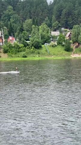 A person is water skiing on a wide river. The person is wearing dark clothing and is in the center left of the frame. They are leaving a white wake behind them. In the background, there is a densely forested hill with several houses scattered among the trees. The sky is overcast. To the right of the water skier, there are two small boats with people in them. The water is choppy.