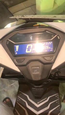 The video shows a close-up of a motorcycle's digital dashboard. The dashboard displays the time as 5:22, the speed as 0 km/h, and a fuel gauge that is nearly full. A person's foot is visible at the bottom of the frame, wearing a sandal. The background is out of focus but appears to be an outdoor environment at night, with some foliage and a building visible in the distance. The motorcycle is white and has a Honda logo. The video is shaky, suggesting it was recorded by someone holding the camera.