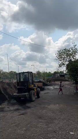 A yellow front-end loader is parked on a dirt road. A person is walking away from the loader. There are piles of dark material on either side of the road. The sky is cloudy.