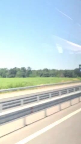 The video shows a view from a moving vehicle, likely a bus or car, traveling on a highway. The scene is dominated by a clear blue sky with faint contrails. On the right side of the road, a billboard with a green background and text that appears to be in Russian or a similar Cyrillic alphabet is visible. The billboard features an image of a man and possibly a soccer ball. Below the billboard, there is a line of trees and foliage. On the left side of the highway, there is a field of green crops, and further back, a dense line of trees. Two cars are visible on the highway. The first car, a dark sedan, is moving in the same direction as the camera. The second car, a white sedan, is also moving in the same direction. The highway has a white lane marker and a guardrail. The overall environment is rural and appears to be a sunny day.