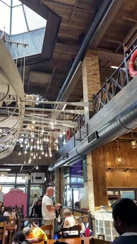 The video shows the interior of a restaurant with a rustic, industrial design. A large, unfinished wooden boat hull is suspended from the ceiling, serving as a prominent decorative element. Numerous bare light bulbs hang from strings, creating a warm and ambient lighting effect throughout the space. The ceiling is a mix of wooden planks and a large skylight, allowing natural light to filter in. Exposed brick walls and wooden beams contribute to the overall aesthetic.

In the foreground and midground, tables with diners are visible. People are seated, eating, and conversing. Some staff members, dressed in casual attire, can be seen moving around the restaurant, possibly serving or attending to customers. In the background, there are various food stalls or service counters, suggesting a market-style or food court setup within the restaurant. The atmosphere appears lively and casual, with a mix of natural and artificial light creating a dynamic visual environment.