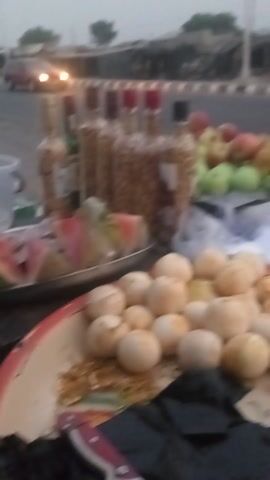 The video displays an outdoor scene, likely a food stall or market stand, filmed with a vertical orientation, causing the contents to appear rotated ninety degrees clockwise.

On the right side of the frame, which would be the bottom if the video were viewed horizontally, a food stall is laden with various items. In the immediate foreground, a round, white tray with a red rim holds several small, pale, round objects, resembling boiled eggs or small mushrooms. Alongside these are a few small, orange, spherical fruits, possibly apricots, and a scattering of shredded orange vegetables, likely carrots. A dark, flat object, possibly a utensil or a piece of food, is also present on this tray.

Behind this tray, another surface or tray exhibits more food. Slices of red watermelon are prominent, along with a large, pale, round root vegetable cut in half. Smaller, light-colored spherical items, which could be onions or other fruits, are also visible. Further back within the stall, there is a large mound of irregularly shaped, brownish items, some of which are contained in clear plastic bags, while others are loose. These appear to be fried snacks or nuts. Below these, some green vegetables, possibly cucumbers, are arranged.

On the far left side of the frame, which would be the top if viewed horizontally, a street scene is depicted. Blurry vehicles, including a red car and a blue vehicle, are visible, suggesting traffic passing by. The general lighting is dim, indicating either an overcast day, late afternoon, or early evening. The accompanying audio features background chatter and ambient street noise, suggesting a bustling environment. No specific human actions are clearly discernible, and no text is visible.