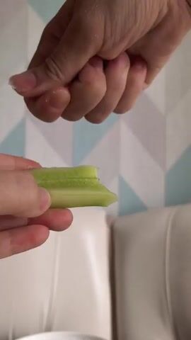 A close-up, vertical shot shows two hands engaged in preparing food. In the foreground, a person's left hand holds a segment of green celery. From above, another person's right hand sprinkles granular white sugar onto the celery. The background features a textured wallpaper with a geometric pattern of grey and teal rhombuses and a white, plush surface, possibly upholstery. The lighting is soft and natural.