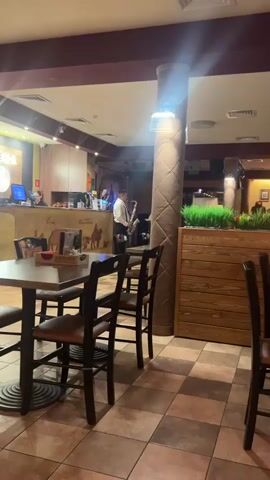 The video shows an indoor scene of a restaurant. A man in a white shirt and black pants is playing a saxophone in the middle of the restaurant. There are tables and chairs visible in the foreground, suggesting a dining area. In the background, there is a counter or bar area with people working. The restaurant appears to have a warm and somewhat dimly lit ambiance, with checkered floor tiles and decorative pillars. There are also plants and wooden accents adding to the decor. The focus is on the saxophonist performing for the patrons or ambiance of the establishment.