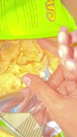 The video shows a close-up view of a person's hands reaching into a bag of potato chips. The hands are picking up a chip from the bag, which is partially visible at the top of the frame. The chips appear to be thin and crispy.

Visible text on the bag includes ingredients and nutritional information. Some of the text is in Indonesian, and some is in English. One prominent phrase in Indonesian reads, "Kelapa Sawit, Bumbu Penguat Rasa Mononatriu," which translates to "Palm Oil, Flavor Enhancer Monosodium Glutamate." Other visible ingredients include "Bubuk Susu," which means "Milk Powder," and "Sour Cream (0.55%)." There is also a mention of "Kota Pematang Siantar, Indonesia," indicating the location.

The lighting suggests it is daytime, possibly indoors. The focus is on the hands and the chips, creating a sense of casual consumption or preparation. There are no strong emotional cues, but the action is straightforward and implies the person is about to eat a chip. The background is blurred but hints at a patterned surface, possibly a table or placemat.