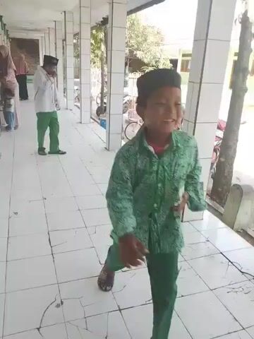 The video shows a tiled corridor with white pillars. A man in a gray shirt and hat sits on the edge of the corridor with his feet dangling outside. Two young boys stand near him. One is wearing a green shirt and the other is wearing white. People can be seen in the background, some are standing and some are walking. Outside, there are bicycles and motorcycles parked. There are also trees and houses visible. The camera is moving slightly.