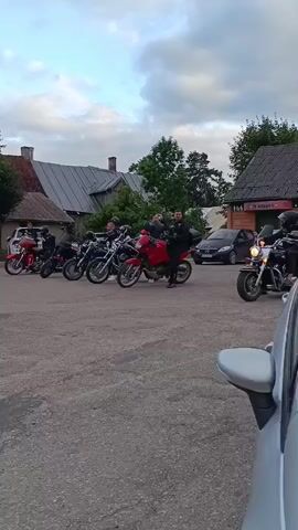 A group of motorcyclists are gathered in a parking lot, some on their bikes and others standing. The motorcycles are a mix of styles, including cruisers and a red sportbike. The people are dressed in motorcycle gear, including helmets and leather jackets. The environment appears to be a rural or semi-rural setting, with buildings and trees in the background. The sky is cloudy, suggesting it may be late afternoon or early evening.