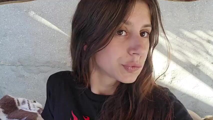 This is a close-up video of a young woman with long, dark, wavy hair and brown eyes. She is wearing a black t-shirt. The background appears to be an outdoor setting with a textured wall and a brown canopy overhead. There are shadows from sunlight casting patterns on the wall. The woman is looking directly at the camera and appears to be speaking or posing. She briefly raises her hand to her hair.