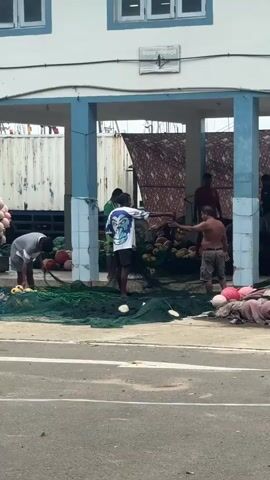 The video depicts a group of people in an outdoor market or fishing area. In the foreground, a person wearing a grey shirt and dark pants is kneeling and appears to be working with a large green fishing net spread out on the ground. To the right of this person, another individual in a white t-shirt with a graphic design and dark shorts is also standing near the net. Behind them, a man wearing a red shirt and dark shorts is visible. Further back, more people are present, and there are piles of what appear to be fishing floats or buoys in various colors, including red and pink. Fishing nets are a prominent feature of the scene. The environment suggests a coastal or harbor setting, with a building with blue columns and a white facade in the background. The ground is paved, and there is a sense of ongoing activity related to fishing equipment.