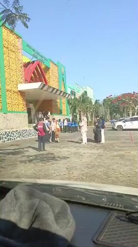 The video shows an outdoor scene in front of a building with green and yellow facade. Several people are standing around in the paved area. In the foreground, a person in a red vest and black pants is facing away from the camera. To their right, several people are gathered, some wearing traditional Islamic clothing. In the background, more people are visible, and a white car is parked. The sky is clear and blue, suggesting a sunny day. The overall impression is that of a gathering or event taking place outside the building.