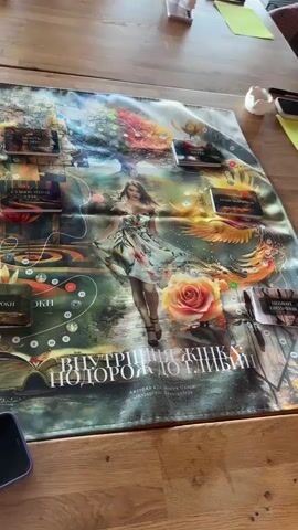 Here is a description of the video:
The video shows a group of people sitting at a wooden table playing a board game. The board game has an artistic design featuring a woman, a rose, and a phoenix. The game's name is "Внутрішня подорож до глібін" (Internal Journey to the Depths). There are cards and markers placed around the board. Each person has a yellow piece of paper and a pencil in front of them. The people appear to be engaged in the game, looking at the board and discussing. Large windows behind them provide a view of a green landscape. There is a phone on the table near the game.