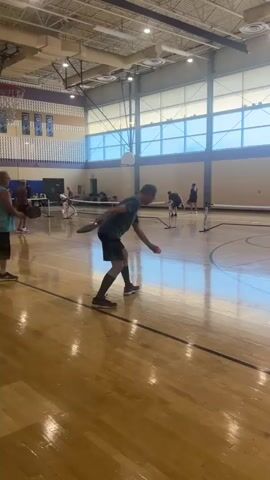 This is a vertical, full-shot video filmed inside a gymnasium. The floor is a polished wood gymnasium floor. A pickleball net is set up in the middle of the court. There are several people playing pickleball. In the foreground, a man in a dark green shirt and black shorts is facing away from the camera and is in the process of hitting a pickleball. He is wearing dark socks and athletic shoes. In the middle ground, a man in a teal tank top and black shorts is also playing pickleball and is facing the camera. In the background, there are more people playing pickleball. The gymnasium has a high ceiling with fluorescent lighting. Large windows line one wall of the gymnasium, letting in natural light. There is a basketball hoop visible on the left side of the frame. The overall environment appears to be a recreational center or school gymnasium.