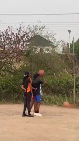 Three individuals stand in an outdoor setting. The central figure, dressed in a black and blue striped shirt and blue shorts, is about to shoot a basketball. To their left, a person in a black shirt with orange accents and black pants watches. On the right, another person in a black shirt and pink shorts observes the basketball shot. A building and dense greenery are visible in the background. A tall pole stands nearby, adding to the outdoor atmosphere.