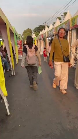 The video shows a street scene with several stalls set up along a paved walkway. The stalls are white tents with various signs and displays. People are walking along the walkway, browsing the stalls. In the foreground, a woman with glasses and a green shirt is walking towards the camera. Behind her, a person wearing a hijab and carrying a pink backpack is walking away from the camera. To their right, a child in red is standing with an adult. Further down the walkway, a young boy in a white t-shirt is standing near a stall. The lighting suggests it is late afternoon or early evening, with the sun casting long shadows. The environment appears to be an outdoor market or festival.