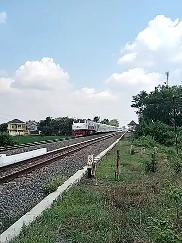 The video shows a train traveling along a railroad track. The train is white with colored stripes. The locomotive is red and white, bearing the KAI logo. The railroad track is lined with gravel and a concrete barrier. A distance marker with the number "3 240" is visible. The environment includes a field of grass and trees. The sky is blue with puffy clouds.