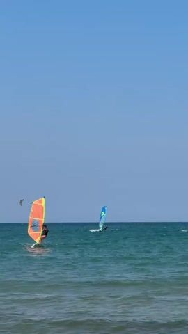 Two windsurfers are visible on a bright, clear day. The windsurfer in the foreground is on the left, with a colorful orange and yellow sail. This windsurfer is actively engaged in the sport, leaning into the wind. Another windsurfer is further out in the water to the right, with a blue sail. The ocean is a deep blue-green, with gentle waves. The sky is a clear, cloudless blue. In the distance, a small object appears to be flying in the sky, possibly a bird or a kite. A few other distant figures are barely discernible on the water. The scene conveys a sense of recreational activity on the water.