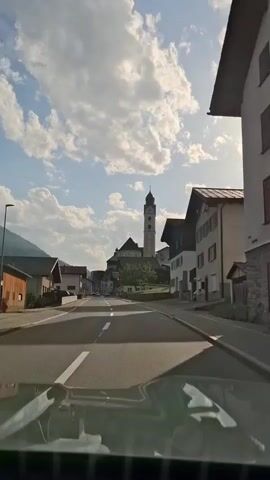 The video shows a view from inside a vehicle, looking forward. The road ahead is paved and has white lane markings. On either side of the road are buildings, typical of a European village. To the left, there is a smaller building with a wooden facade and a grey roof, and further on, a two-story building with white walls and a grey roof. On the right, there are more multi-story buildings with pitched roofs, some with balconies and windows. In the distance, a prominent church tower with a clock face and a spire rises above the other buildings. The sky is blue with scattered white clouds, and the sun is shining brightly, casting shadows. The overall impression is a scenic drive through a picturesque village.