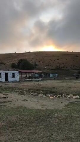 The video shows a rural landscape at sunrise. In the foreground, there is a patch of grass with some puddles of water. Behind the grass, there is a small white building with a red roof, and next to it, a covered structure with a red roof. In the background, a grassy hill rises, and the sun is visible on the horizon behind a cloudy sky. The scene appears to be deserted.