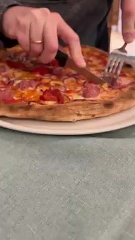 Here is a description of the video:
A close-up shot shows a person cutting a pizza with a knife and fork. The pizza has a thick crust and is topped with cheese, tomato sauce, sausage, red onions, and peppers. The person is wearing a gray long-sleeved shirt and a wedding band on their left ring finger. The pizza is on a white plate, which sits on a blue and white patterned tablecloth. The background is slightly blurred but appears to be an indoor setting.