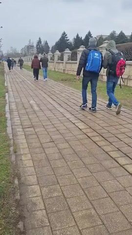 The video captures a street scene in an urban environment. In the initial frame, several people are walking along a sidewalk paved with square stones. Two individuals, one wearing a blue backpack and the other a red one, are prominent in the foreground. As the camera pans, a wide road with multiple lanes comes into view, separated from the sidewalk by a strip of grass and dirt. Several cars are visible on the road, including a yellow taxi.  A large, ornate building dominates the background across the road. The weather appears overcast.  A street lamp is also visible, along with some road construction markers.