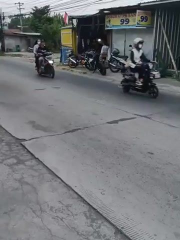 The video captures a street scene with several vehicles and pedestrians. In the foreground, a gray concrete surface with a textured pattern runs horizontally. In the background, a paved road is visible. Several motorcycles are parked along the side of the road next to a building with a blue and yellow awning. The building appears to be a small shop or market, with signs advertising prices. A black car drives down the road, followed by two motorcycles, one red and one black. A person on a motorcycle with a red body and a black seat is riding towards the camera, and then another motorcycle with a black body and a yellow shirt rider is also seen. Another motorcycle with a person wearing a dark jacket and a brown bag is seen in the initial frames. The environment suggests an outdoor commercial or residential area, possibly in a tropical or subtropical climate, given the presence of lush green foliage in the distance. The lighting indicates it is daytime, though the sky appears overcast.