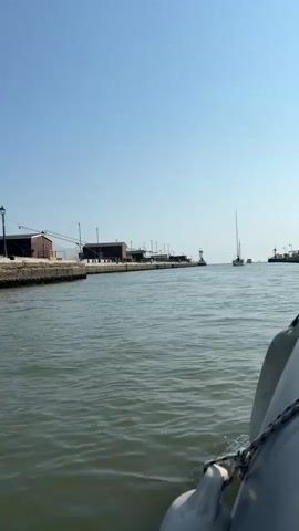 A first-person view from a boat on a body of water. The boat has white fenders on the right side. The water is choppy and green. Across the water, a concrete wall lines a dock with several buildings. In the distance, several sailboats and a lighthouse are visible. The sky is clear and blue. The overall impression is a sunny day in a harbor.