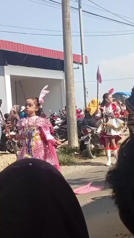 The video shows a street scene with people in parade costumes. In the foreground, a young girl in a pink and gold dress with a feather in her hair stands and looks to her left. To her right, a taller individual in a white and red marching band uniform, complete with a feathered headdress and boots, holds a staff with flags. This person is marching or dancing, with their left leg extended. Behind them, there are several motorcycles parked and people watching. A building with a red roof is visible in the background, along with utility poles and wires. The day appears sunny. The camera is positioned low, with the dark upper portion of a person's head or head covering in the bottom foreground.